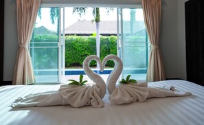 Una cama con dos toallas dispuestas en forma de cisnes, una ventana con cortinas y vistas al exterior con vegetación.