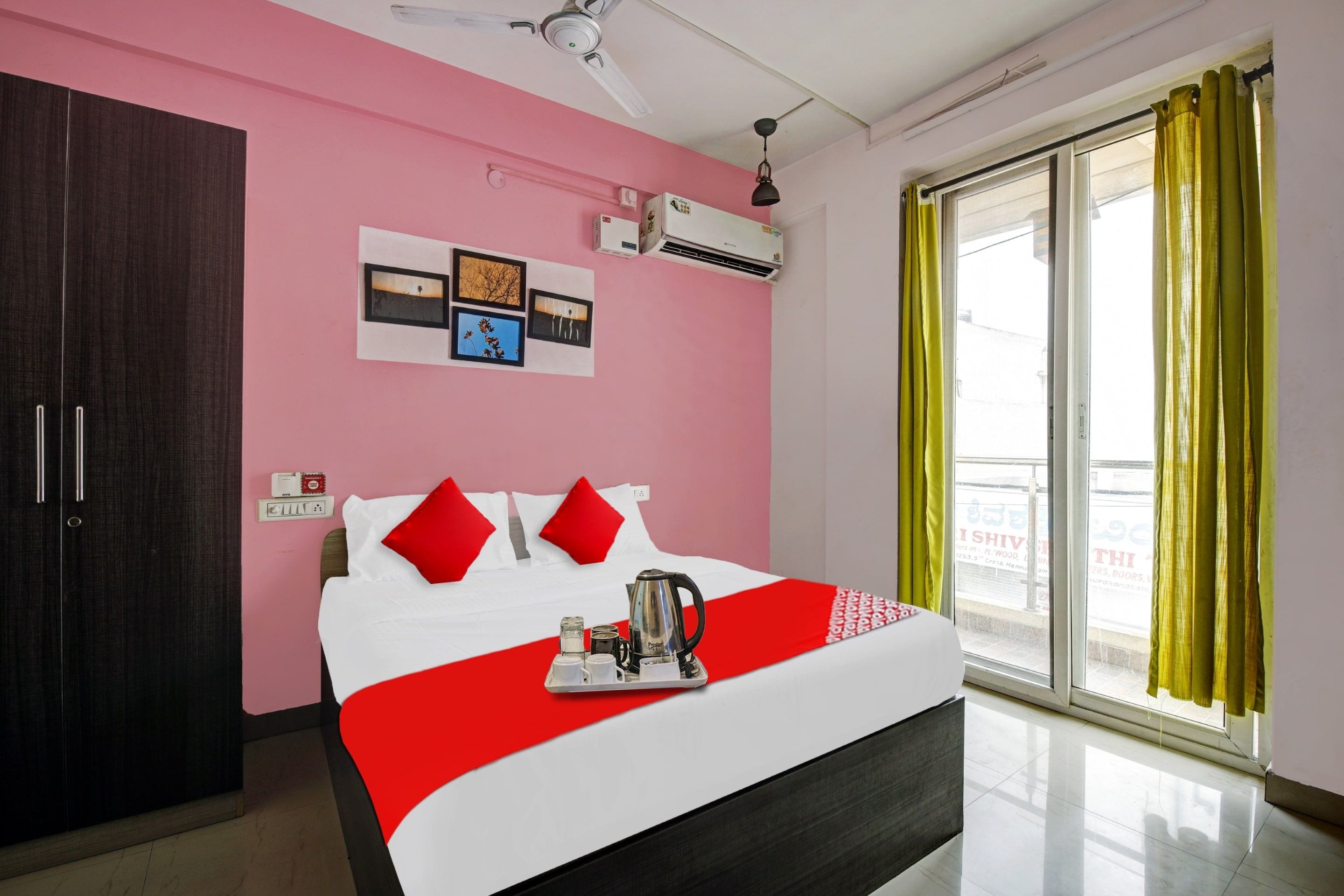 Quarto de hotel com cama, guarda-roupa, ventoinha no teto, ar condicionado, janela com cortinas e parede decorada com quadros.