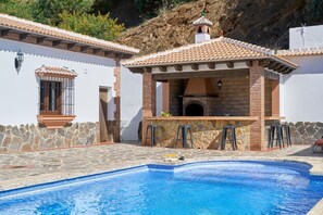 A swimming pool with a stone deck, a barbecue area with stone walls, and a small window with a view of the surrounding landscape. - Villa Alejandro in Sayalonga. Located on a quiet, sunny private estate (Sayalonga)