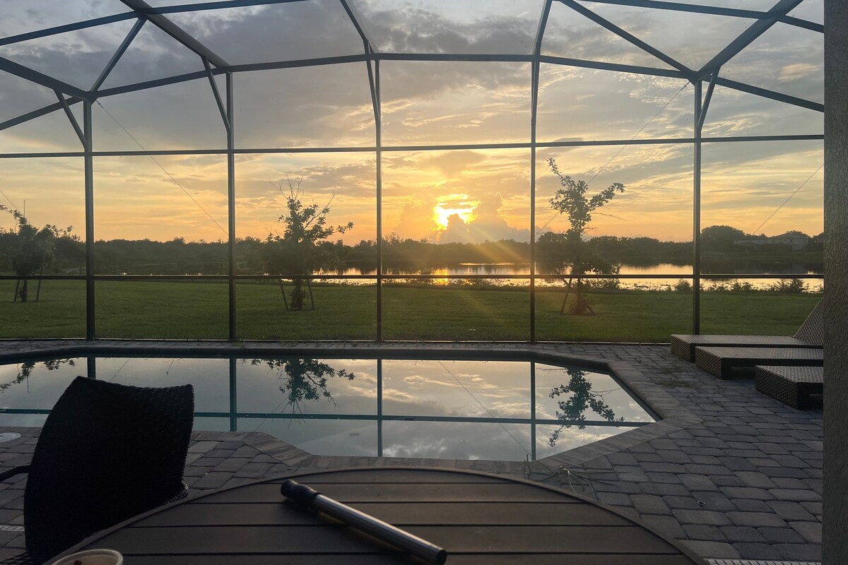 A glass-enclosed outdoor area with a view of a sunset over a lake, a pool, and a patio with seating.