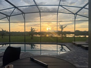 Area esterna vetrata con vista su un tramonto sul lago, una piscina e un patio con posti a sedere.