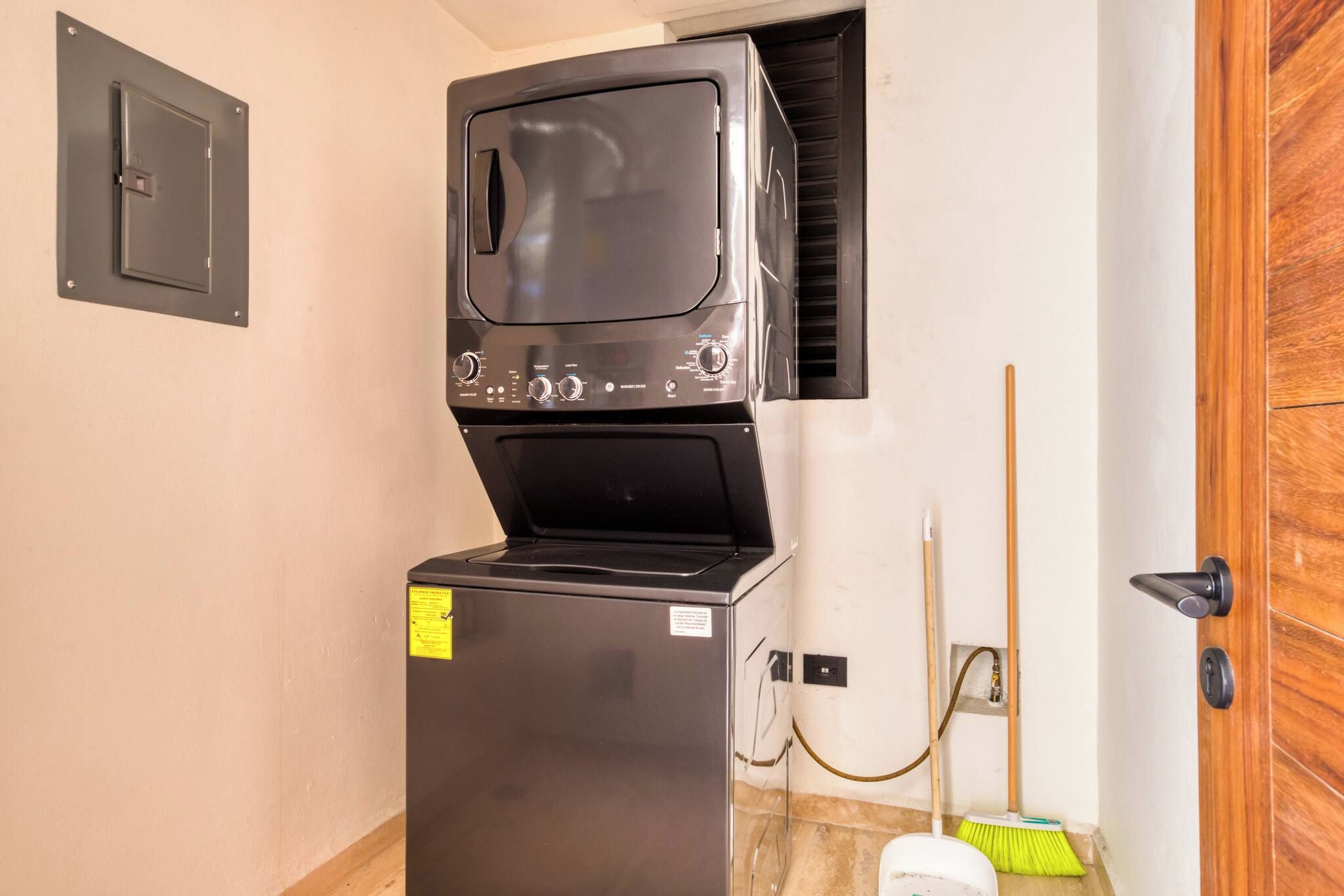 Una unidad compacta de lavadora y secadora instalada en una habitación pequeña con una puerta de madera y una escoba apoyada contra la pared.