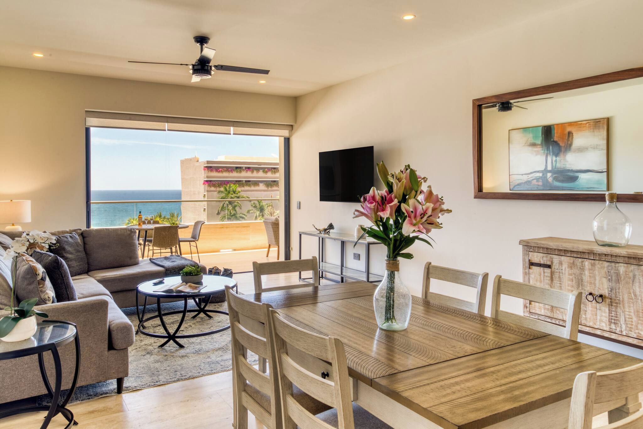 A spacious living area with a balcony overlooking the sea, a dining table, and a TV mounted on the wall.