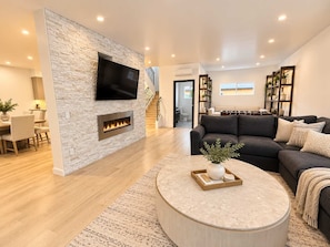 A modern living room with a stone accent wall, a flat-screen TV, a fireplace, a dark sofa, and a round coffee table with decorative items. - Point Loma Sleeps 16 | Rooftop | Beach & Downtown (San Diego)