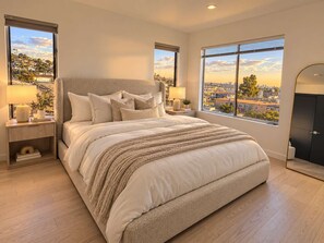 A modern bedroom with a large bed, two bedside tables, and a view of a cityscape through large windows. - Point Loma Sleeps 16 | Rooftop | Beach & Downtown (San Diego)