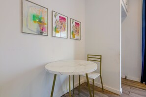 A room with a round table and a chair, a wall with three abstract paintings, and a blue curtain. - Steps to Santa Monica Beach & Pier! Monthly Studio (Santa Monica)