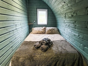 Ein schmales Zimmer mit schrägem Dach, einem Bett mit zusammengerollten Decken und einem Fenster mit Blick ins Freie.