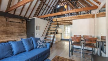 Ein Loft-Zimmer mit freiliegenden Holzbalken, einem blauen Sofa, einer Essecke mit Stühlen und einer Treppe, die zu einer erhöhten Ebene führt.