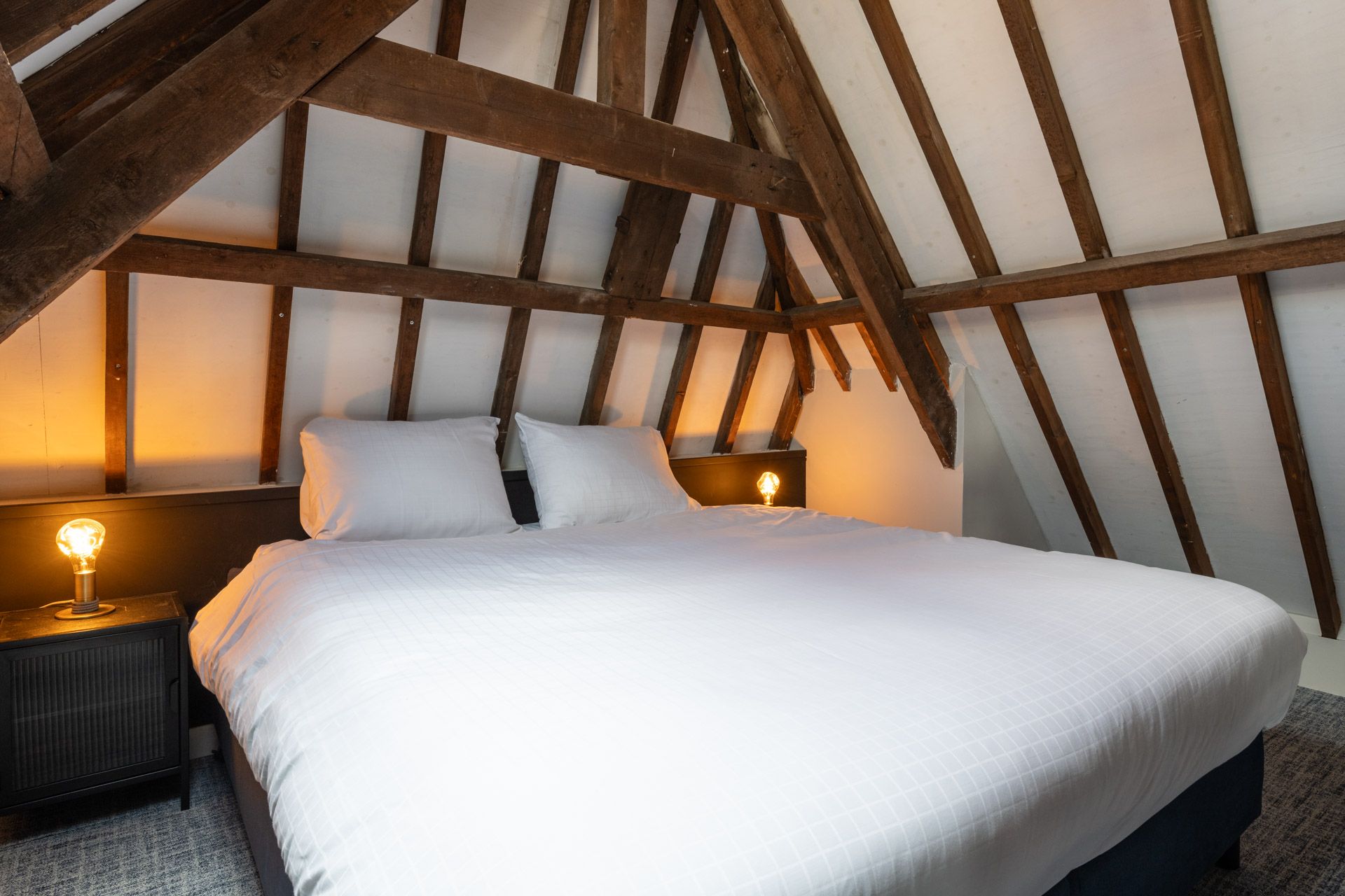 Une chambre avec un plafond en pente, des poutres apparentes, un lit avec une literie blanche et deux lampes de chevet.