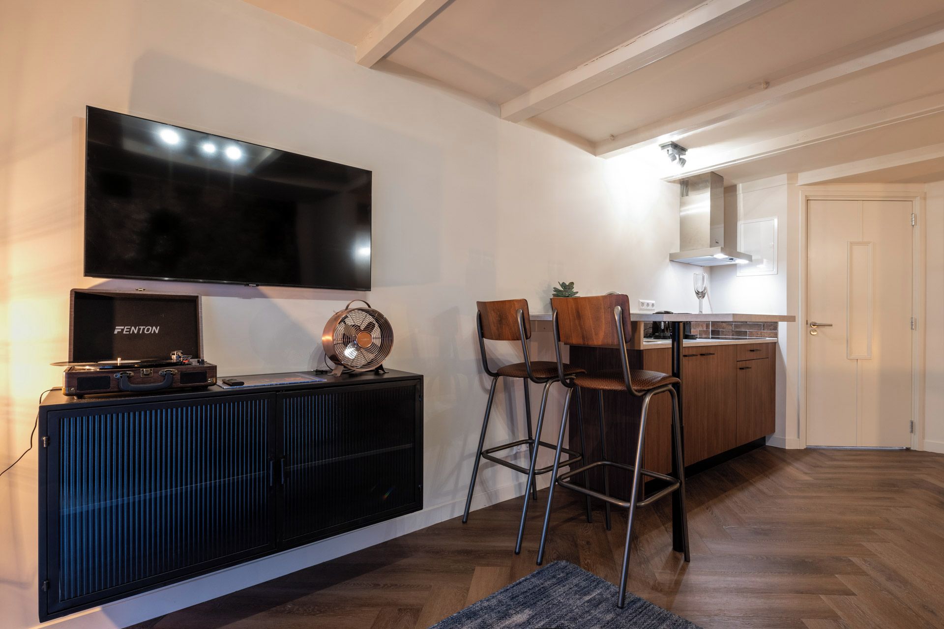 Een moderne woonkamer met een flatscreen televisie, een zwarte entertainmentkast, een barkruk en een keuken met houten kasten en een aanrecht.