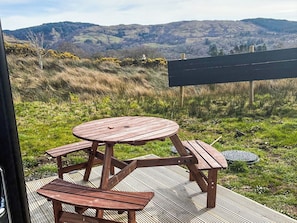 A wooden picnic table with benches on a deck, overlooking a grassy landscape with hills. - 1 bedroom accommodation in Lerags (Lerags)