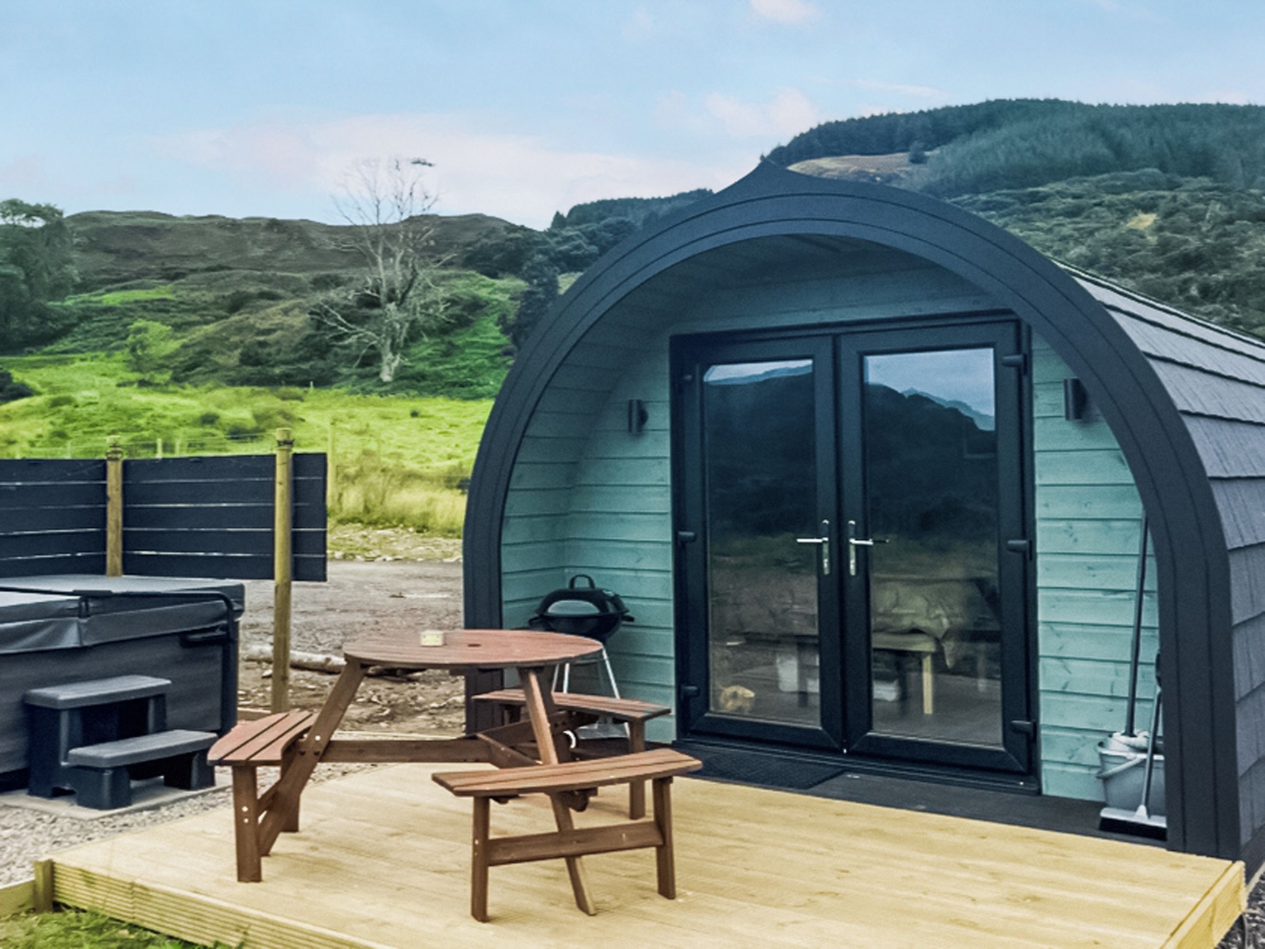 A cozy outdoor seating area with a wooden table and benches, a barbecue grill, and a large arched door leading to a building with a dark exterior.