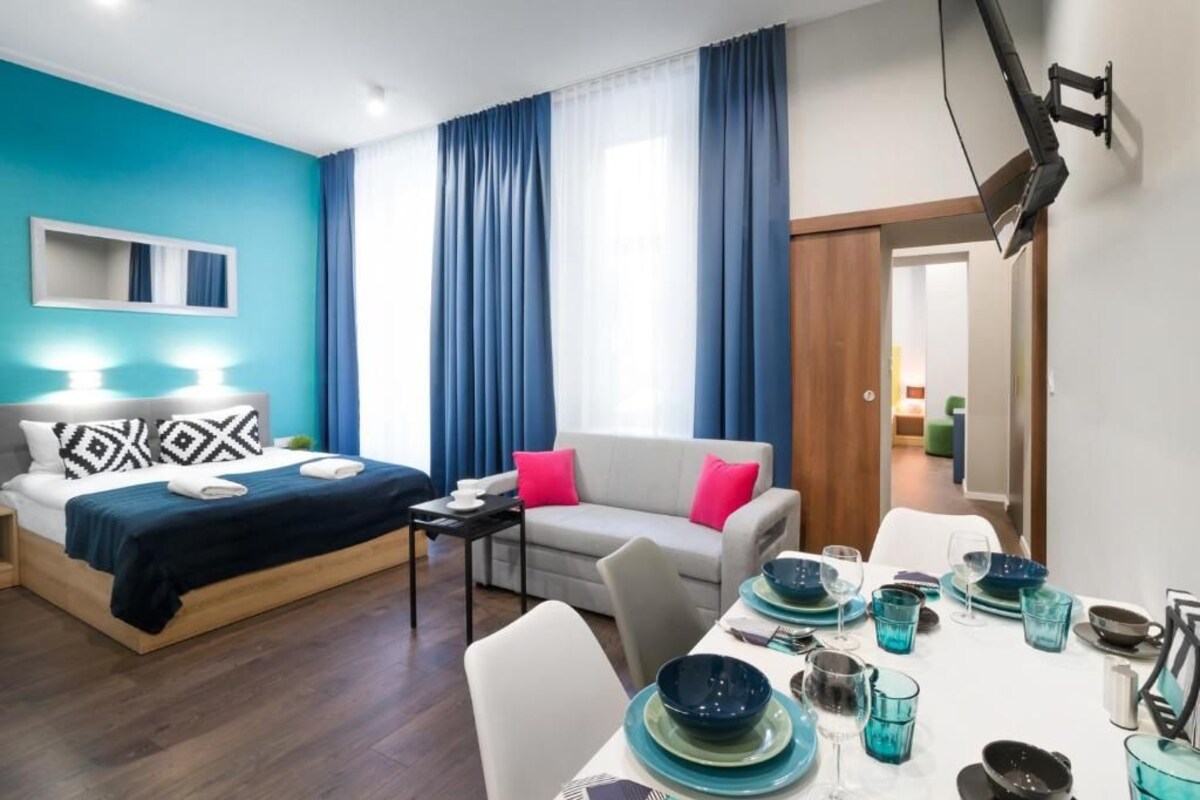 A modern hotel room with a bed, a sofa, a dining table set with blue glassware, and a large window with blue curtains.