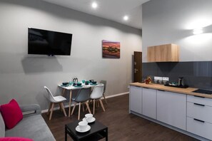 A modern living space with a flat-screen TV, a dining area with chairs and a table, a kitchen with cabinets, and a red pillow on a sofa. - Kings City Sławkowska Apartments (Kraków)