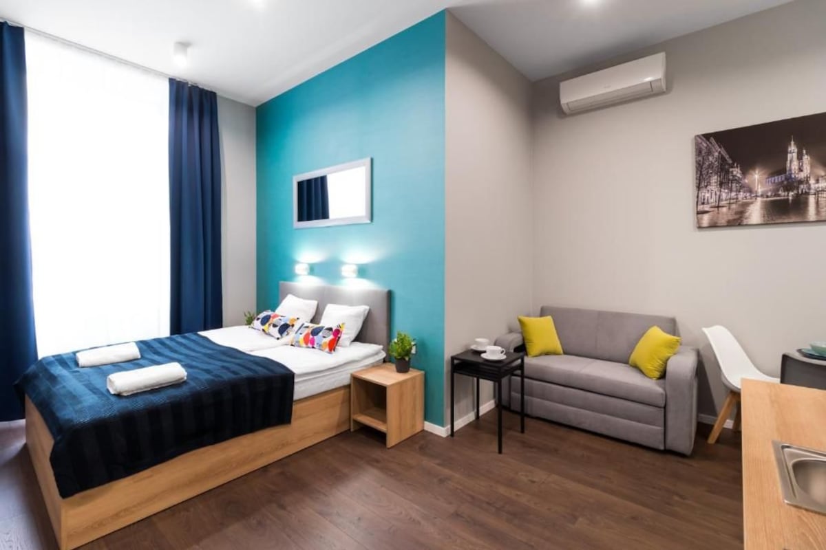 A modern hotel room with a bed, a sofa, a small table, and a wall-mounted air conditioner.