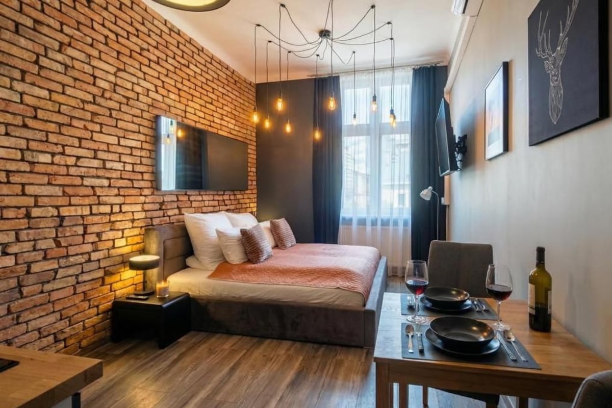 A hotel room with a brick wall, a bed with a headboard, a dining table with a wine bottle, and a chandelier.
