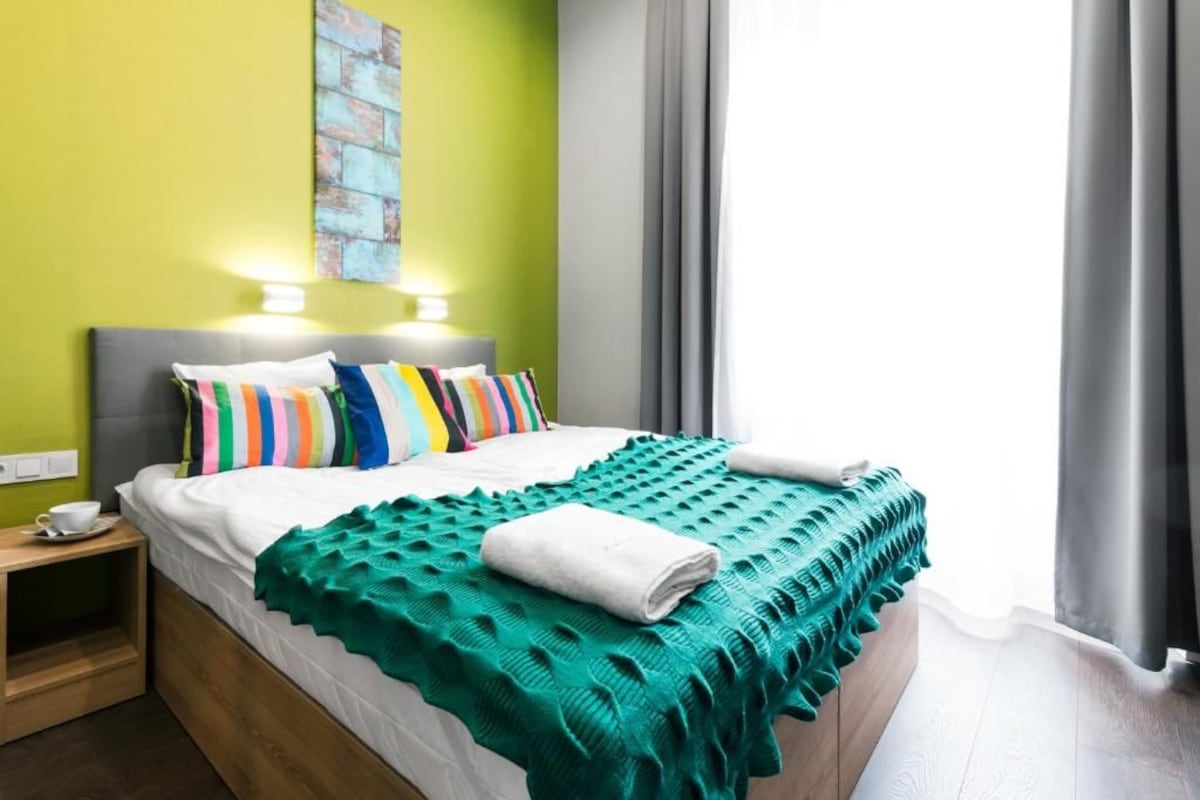 A bed with a white comforter, multiple pillows, a teal blanket, and a bedside table with a cup.
