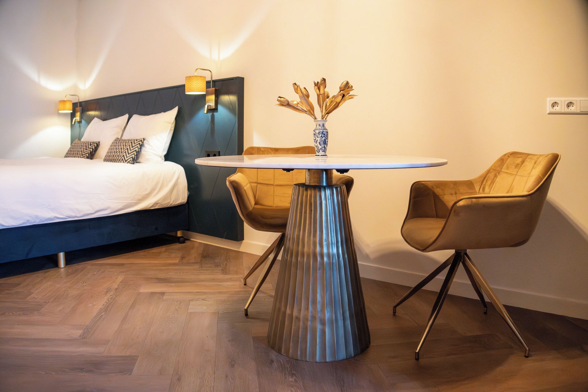 Een hotelkamer met een bed, een ronde tafel met een vaas, twee stoelen en een wandlamp.