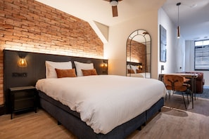 Un dormitorio moderno con pared de ladrillo decorativa, una cama grande con ropa de cama blanca, un escritorio con silla y un espejo grande.