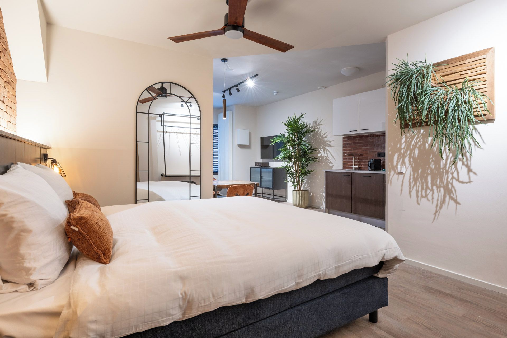 A modern bedroom with a bed, a ceiling fan, a large mirror, and a view into the kitchen area.