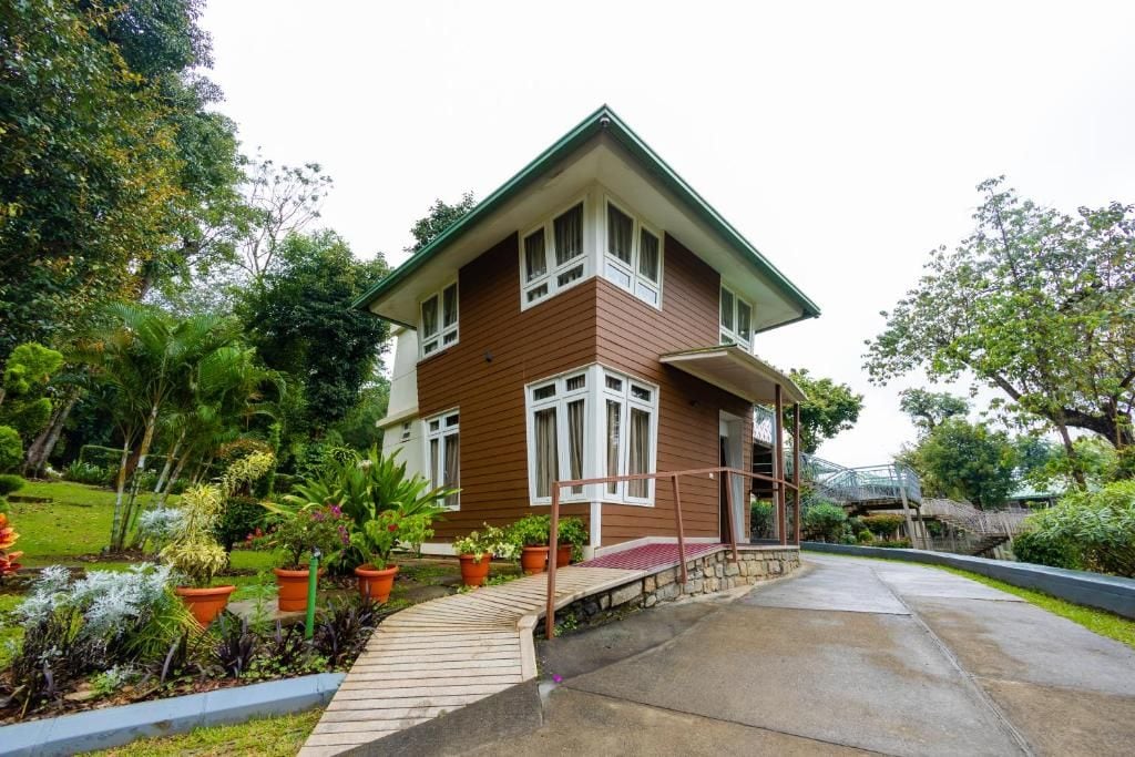 A two-story house with a sloped roof, surrounded by a garden with various plants and a small bridge.