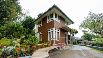 A two-story house with a sloped roof, surrounded by a garden with various plants and a small bridge.