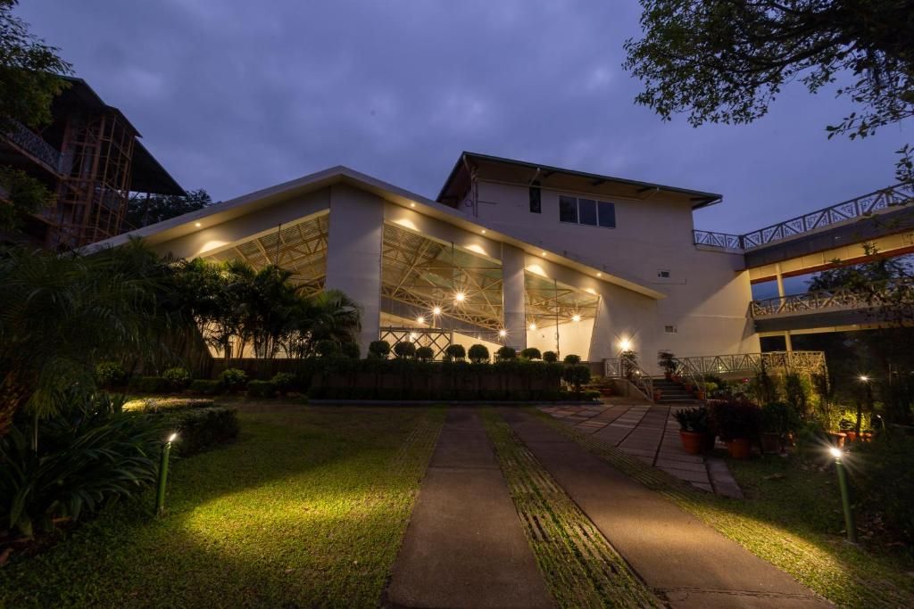 A modern building with a covered outdoor area, surrounded by well-maintained greenery and a pathway leading to the entrance.