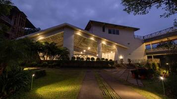 A modern building with a covered outdoor area, surrounded by well-maintained greenery and a pathway leading to the entrance.