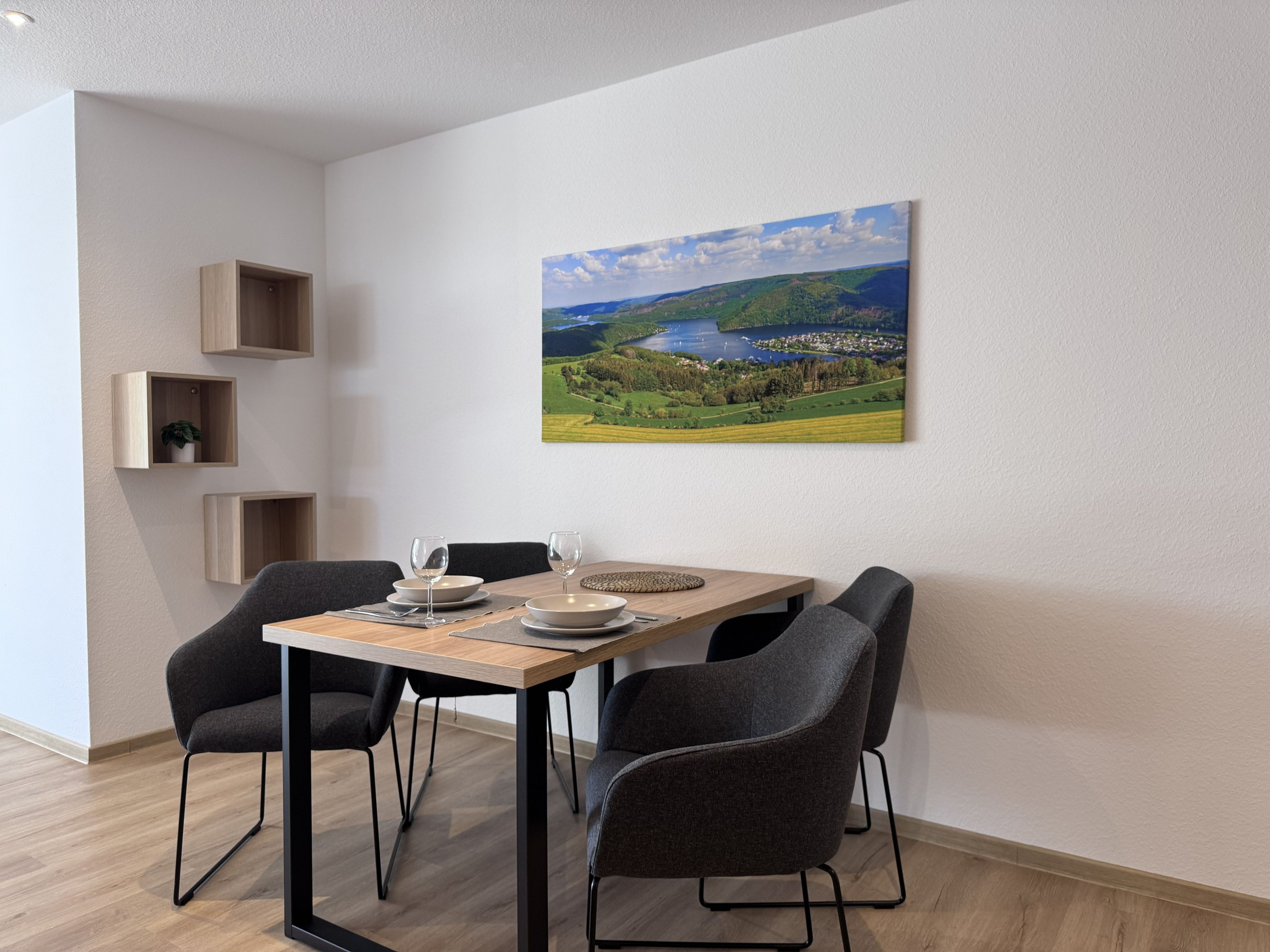 A dining area with a table set for two, two chairs, and a painting of a landscape on the wall.