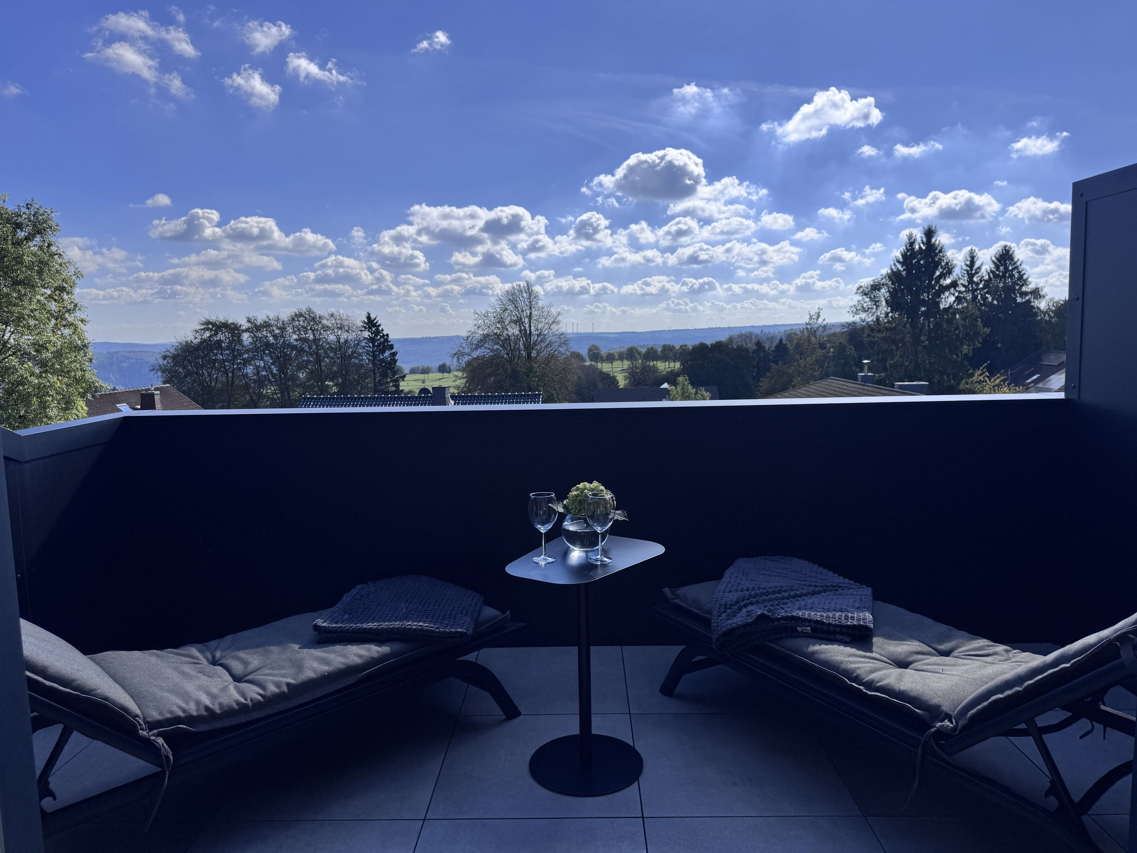 A balcony with two lounge chairs, a small table with a glass and a flower arrangement, and a view of a landscape with trees and a distant hill.