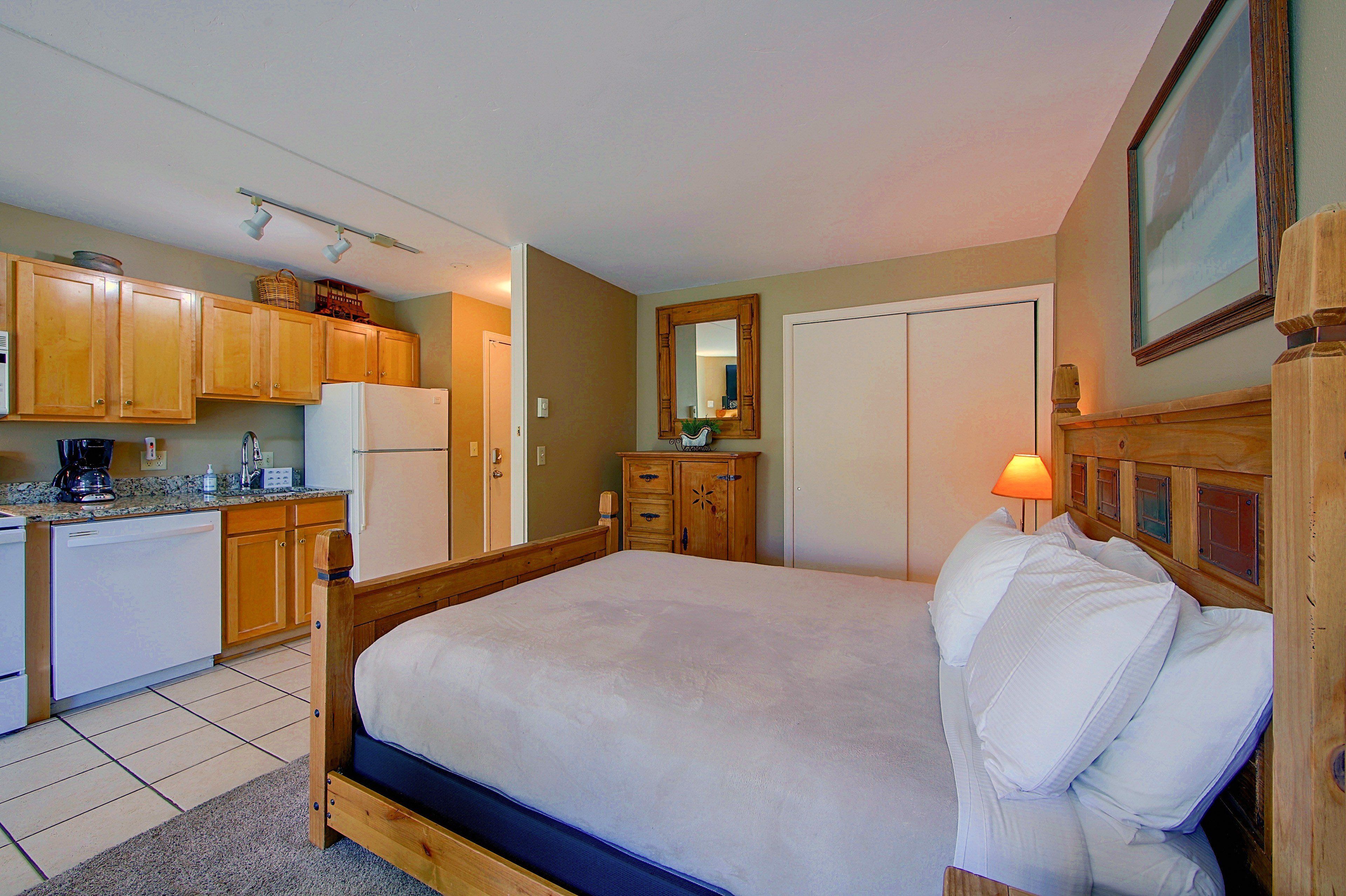A bedroom with a bed, wooden headboard, a dresser, a mirror, and a kitchen area with a refrigerator and cabinets.