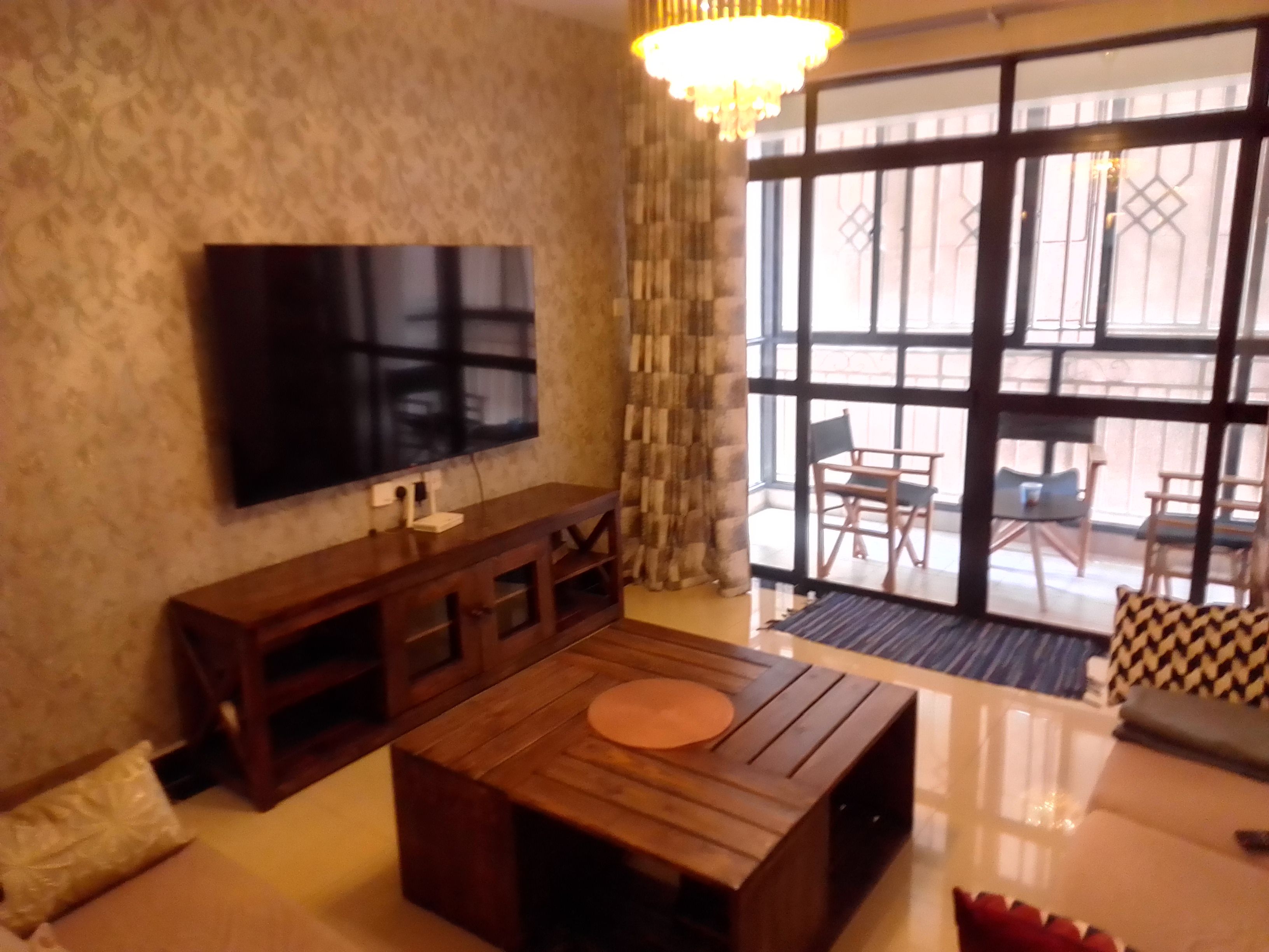 A living room with a flat-screen TV mounted on the wall, a wooden coffee table, a sofa, and a view through large windows to the outside.