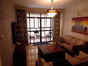 A living room with a sofa, a coffee table, and a television. There is a large window with curtains and a painting on the wall. - Marvelous 4-bedroom apartment in peaceful Nairobi, Kileleshwa neighborhood (Nairobi)
