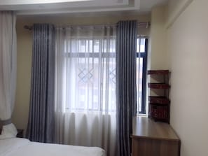 A hotel room with a bed, a window with curtains, and a wooden nightstand with a basket. - Marvelous 4-bedroom apartment in peaceful Nairobi, Kileleshwa neighborhood (Nairobi)