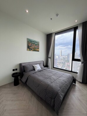 A modern bedroom with a large bed, a painting on the wall, a side table, and a view of the city through a large window. - Panoramic River View Condo, Long Stay (Khlong San)