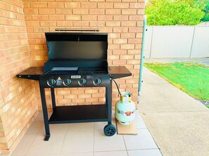 A portable barbecue grill with a gas tank and control knobs, positioned against a brick wall outdoors. - Spacious 3BR, 2 Bath, Dbl garage, WIFI, Step-free (Athol Park)