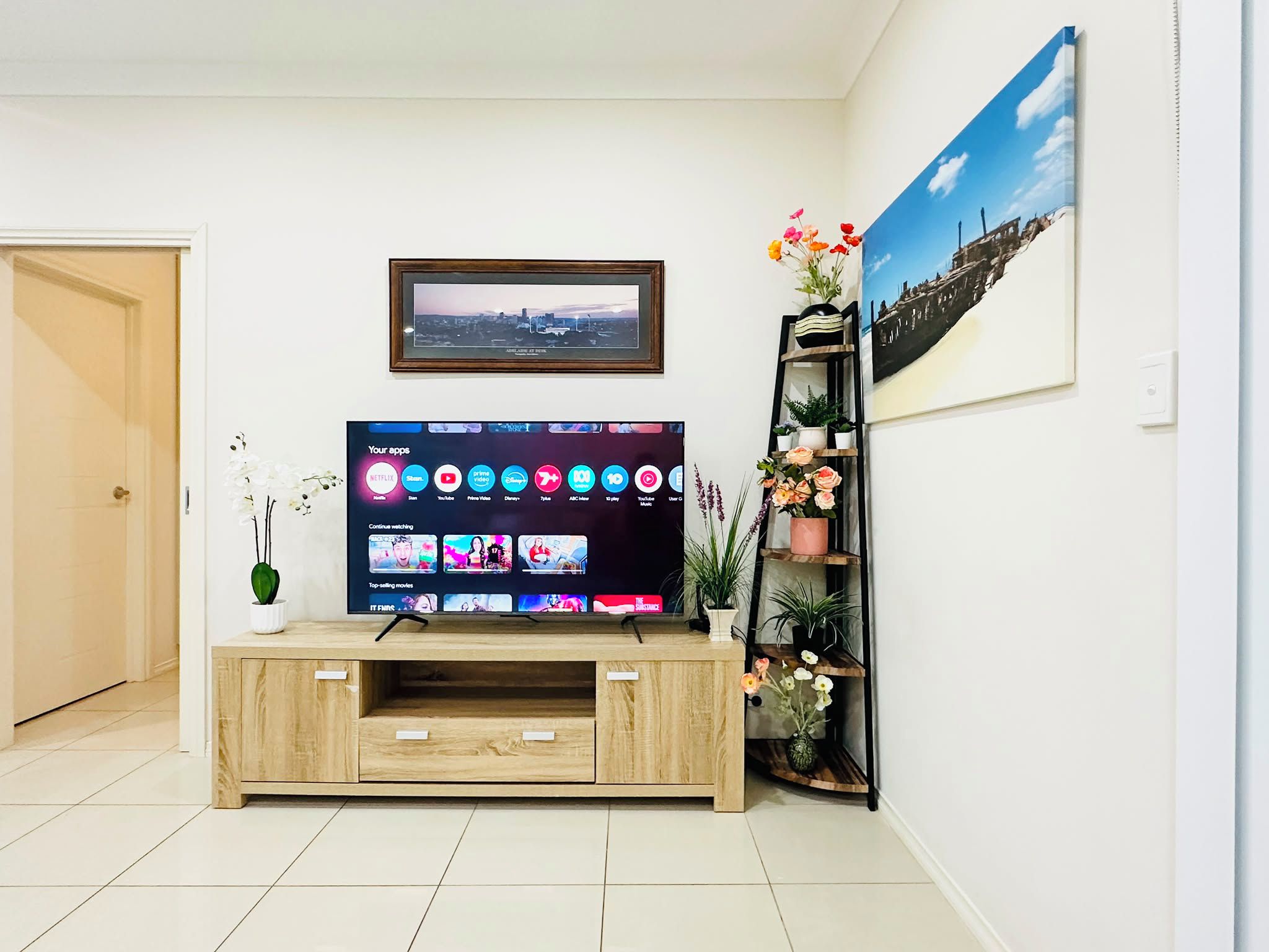 A living room with a flat-screen TV mounted on a wooden entertainment center, a framed picture above the TV, and a potted plant on a shelf to the right.
