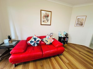 A red leather sofa with patterned cushions, a framed floral artwork on the wall, and a bookshelf with books and decorative items. - Spacious 3BR, 2 Bath, Dbl garage, WIFI, Step-free (Athol Park)