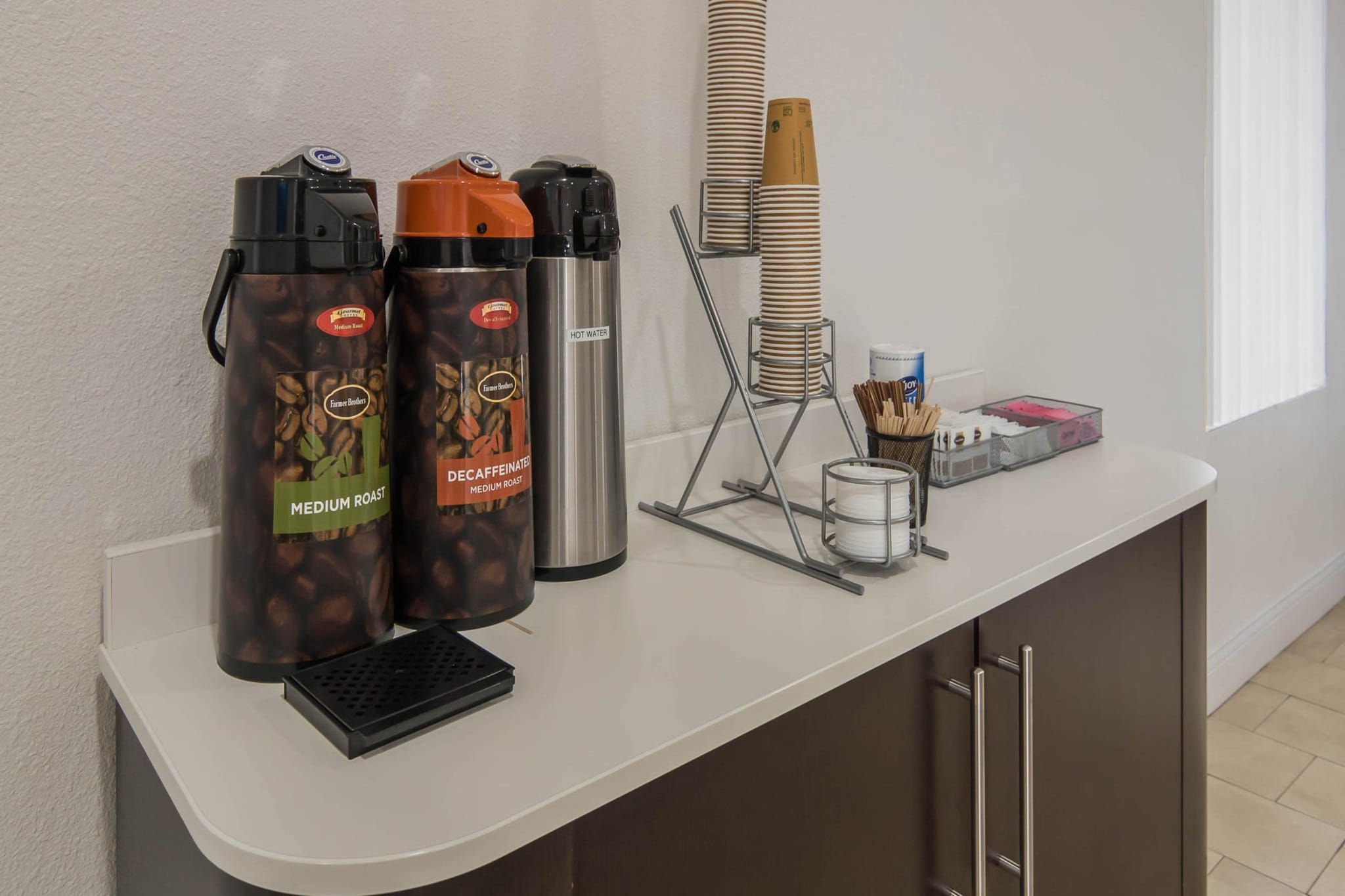 A counter with two coffee dispensers, a hot water dispenser, and a stack of disposable cups.