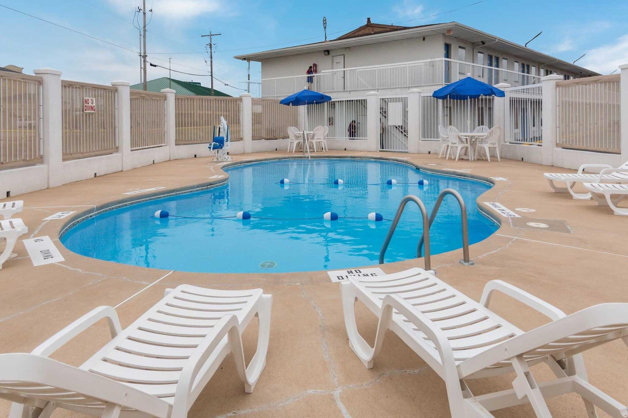 A swimming pool with white lounge chairs and blue umbrellas, surrounded by a white fence with a 'NO DIVING' sign.