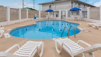 A swimming pool with white lounge chairs and blue umbrellas, surrounded by a white fence with a 'NO DIVING' sign.
