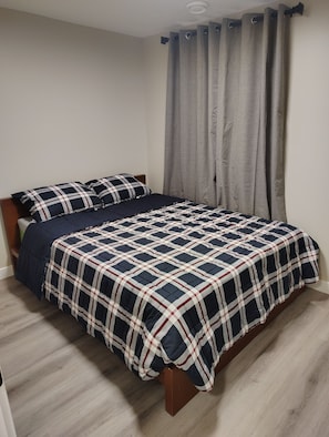 A bed with a plaid bedspread, a wooden headboard, and a grey curtain. - Cozy 3-Bedroom House Rental in Calgary's Rosscarrock Area (Calgary)