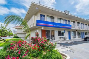 Um motel com um letreiro 'WELCOME', um estacionamento e uma área ajardinada com flores.