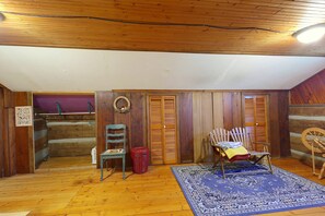 Ein Zimmer mit Holzwänden, einem blauen Teppich mit Muster, einem Holzstuhl, einem Korbwannenstuhl und einer Holztür mit Fensterläden.
