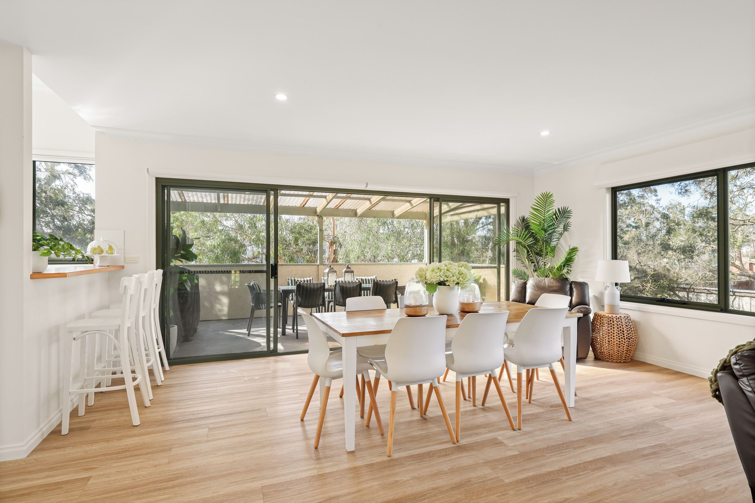 A modern dining area with a wooden table and white chairs, large windows, and a view of the outdoors.