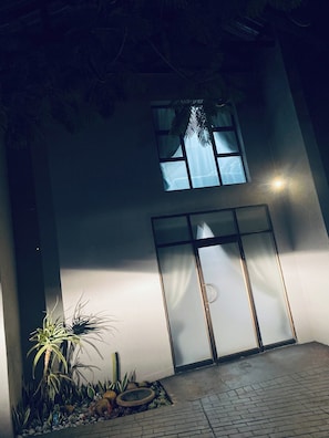 A building with a large window and a door, surrounded by plants and a paved area.