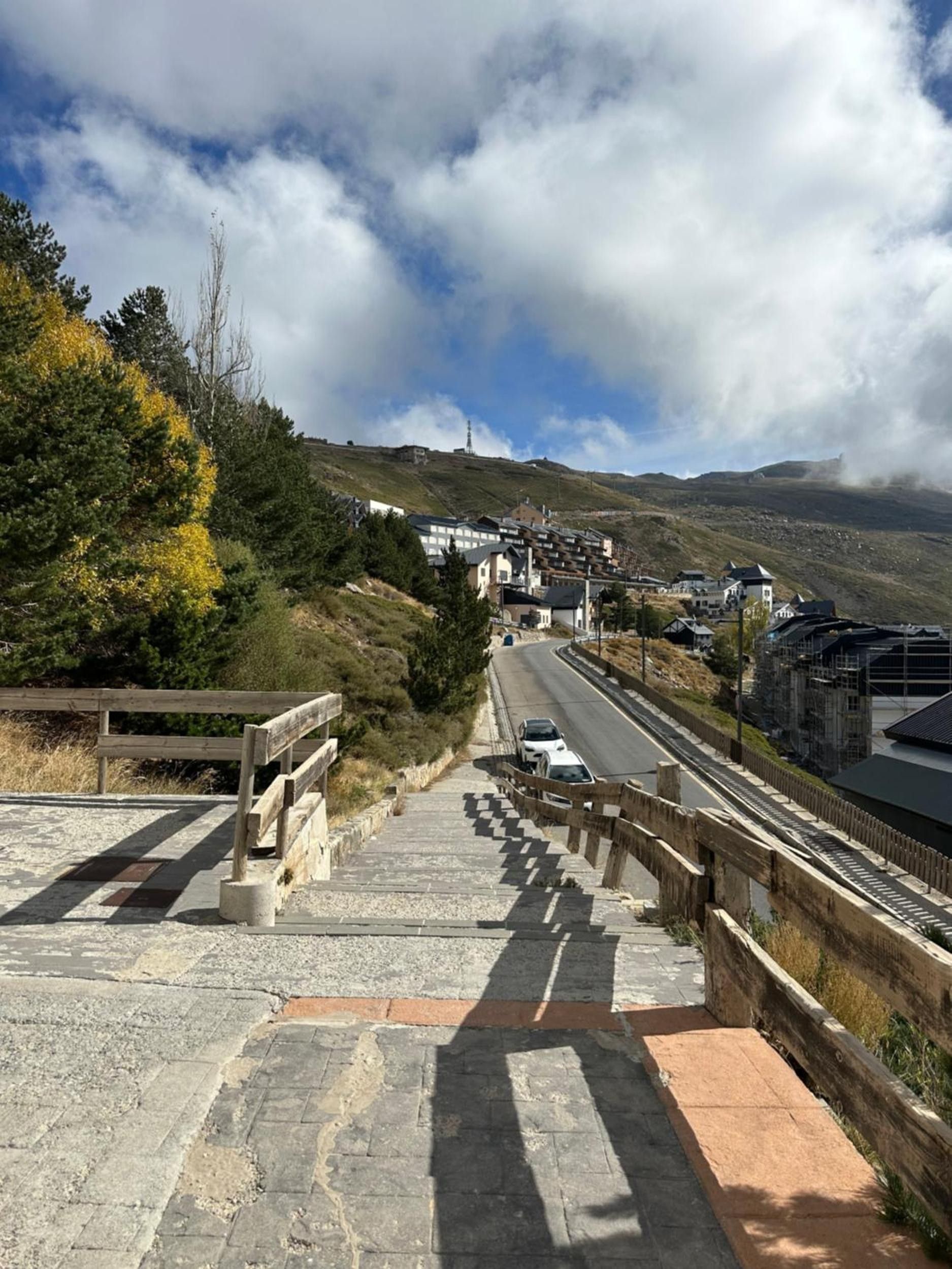 Un camino pavimentado que asciende con una valla de madera a un lado y una carretera al otro.