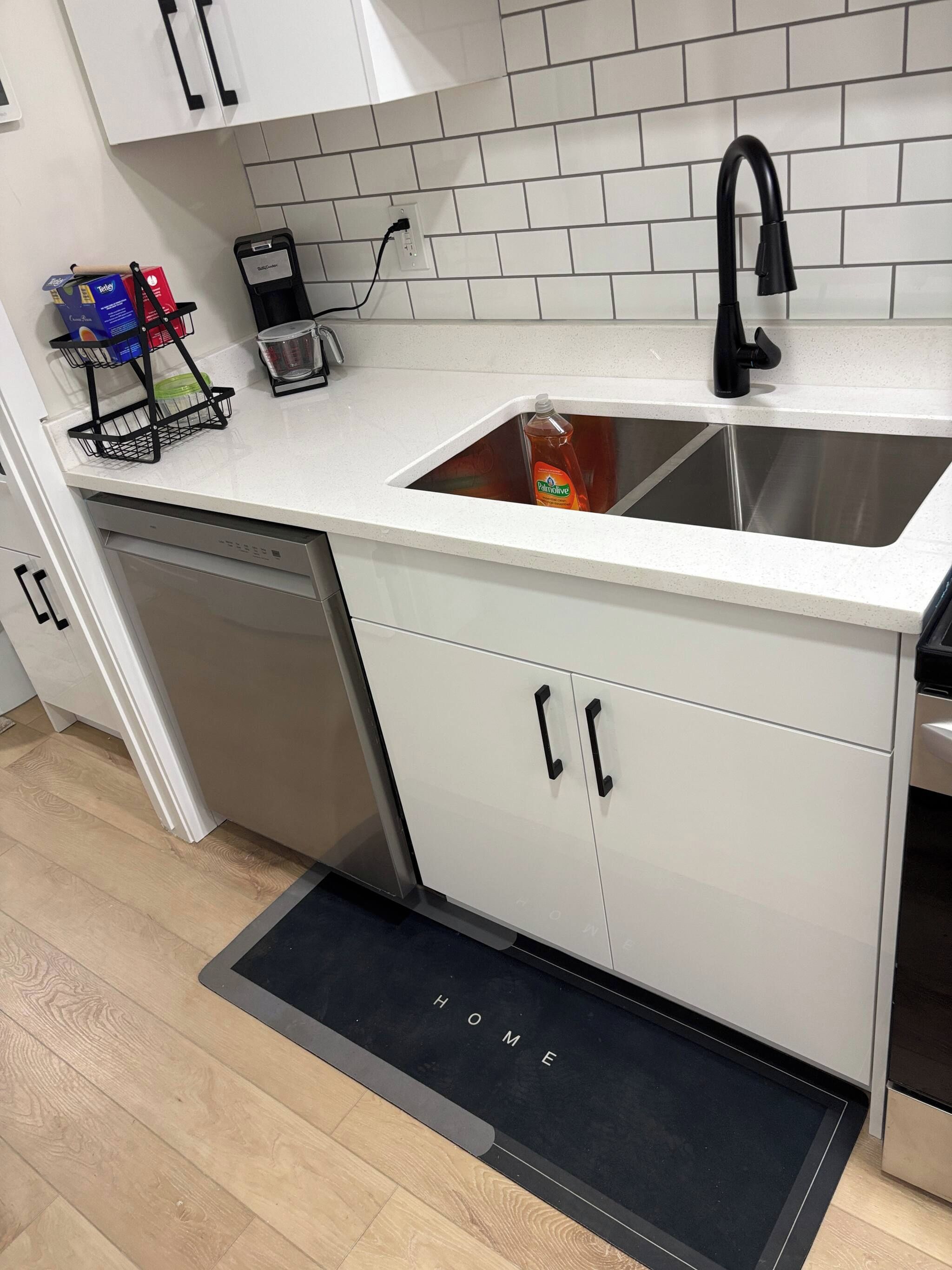 A kitchen with a white countertop, a stainless steel sink, a dish soap bottle, a coffee maker, and a black faucet.