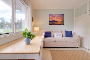 A room with a beige sofa, a blue cushion, a potted plant, a rug, and a sunset painting on the wall.