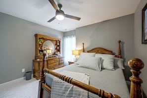 Ein Schlafzimmer mit einem hölzernen Bett, einem Deckenventilator, einer Kommode mit Spiegel und einem Fenster mit Vorhängen.